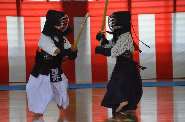 剣道着を着て面を付けた小さい子供2人が、向かい合って打込竹刀を上げている写真